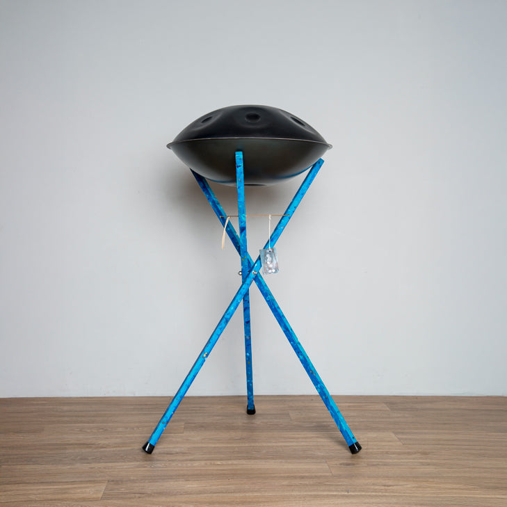 Ein blauer Art of Impro Handpanständer zum in stehen spielen aus Deutschland steht vir einer grauen Wand. Auf ihm liegt eine schwarze Handpan