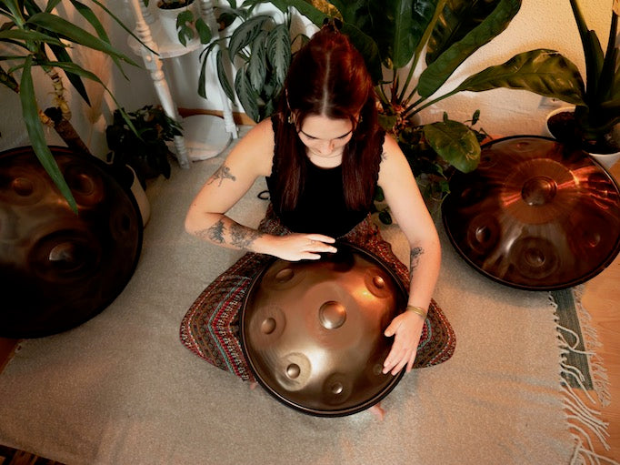 Versierte Handpanspielerin sitzt in einem schönen Raum mit Pflanzen auf dem Boden und spielt eine handgefertigte AeloPan Mini-Handpan mit 9 Tonfeldern aus Edelstahl