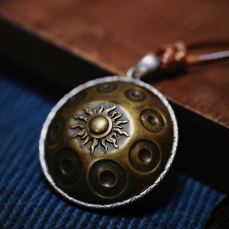 Detailaufnahme einer hochwertigen Halskette mit einem messingfarbenen Handpan-Anhänger. Der runde Anhänger zeigt mehrere Tonfelder wie bei einer echten Handpan, in der Mitte befindet sich ein fein gearbeitetes Sonnensymbol. Der Anhänger liegt auf einem dunklen Stoff und ist an einem Lederband befestigt, was dem Schmuckstück eine edle und handgefertigte Anmutung verleiht.