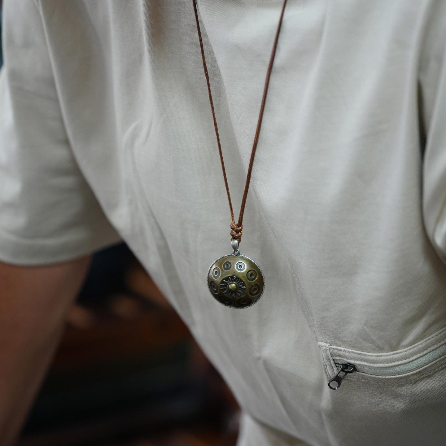 Ein Mann trägt eine hochwertige Halskette mit einem messingfarbenen Handpan-Anhänger, der auf Brusthöhe hängt. Der runde Anhänger zeigt die typischen Tonfelder einer Handpan sowie ein zentrales Sonnensymbol. Die Kette ist an einem braunen Lederband befestigt und wird über einem hellen Shirt getragen, wodurch der Anhänger dezent, aber klar im Fokus steht.