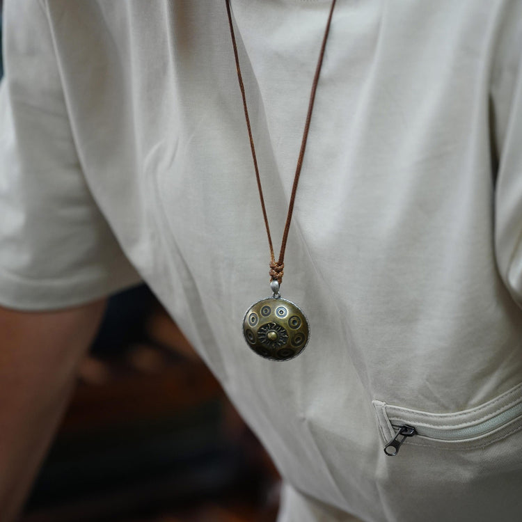 Ein Mann trägt eine hochwertige Halskette mit einem messingfarbenen Handpan-Anhänger, der auf Brusthöhe hängt. Der runde Anhänger zeigt die typischen Tonfelder einer Handpan sowie ein zentrales Sonnensymbol. Die Kette ist an einem braunen Lederband befestigt und wird über einem hellen Shirt getragen, wodurch der Anhänger dezent, aber klar im Fokus steht.