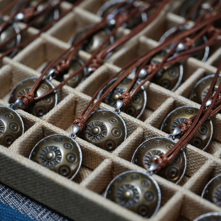 Eine Box mit mehreren identischen Halsketten, ordentlich in einzelnen Fächern angeordnet. Jede Kette besteht aus einem braunen Lederband und einem runden, messingfarbenen Handpan-Anhänger mit typischen Tonfeldern und einem fein gearbeiteten Sonnensymbol in der Mitte.