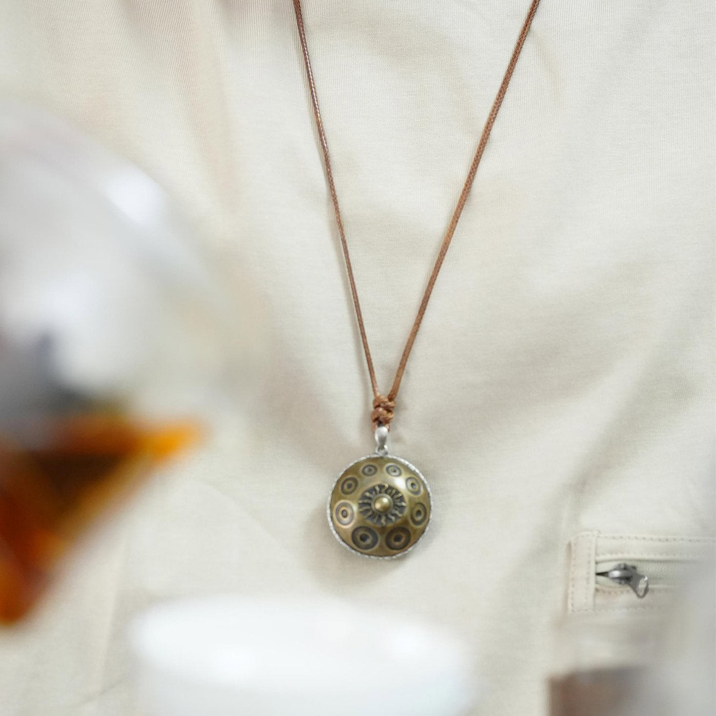 Detailansicht einer hochwertigen Halskette mit rundem, messingfarbenem Handpan-Anhänger, die auf Brusthöhe getragen wird. Der Anhänger zeigt mehrere Tonfelder sowie ein zentrales Sonnensymbol und hängt an einem braunen Lederband. Getragen wird die Kette über einem hellen Oberteil, wodurch der filigrane Schmuck klar hervorsticht und seine handwerkliche Qualität betont wird.