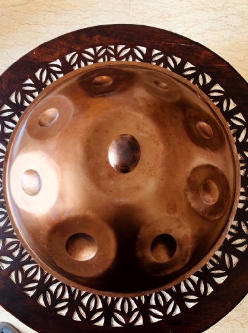 Bronzefarbene Embersteel Svaraa Handpan Drum von oben mit neun Tonfeldern liegt auf einem hölzernen Boden mit Muster. Diese Handpan, sowie viele weitere kann man online bei Handpan.World sicher erwerben.