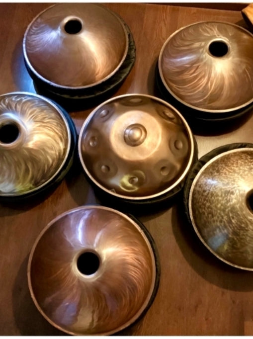 Sechs handgefertigte Svaraa Handpan Drums liegen in einem hellen Wohnzimmer mit weißen Wänden. Sie spiegeln sich im Licht, auf einem ebenen Holzboden.