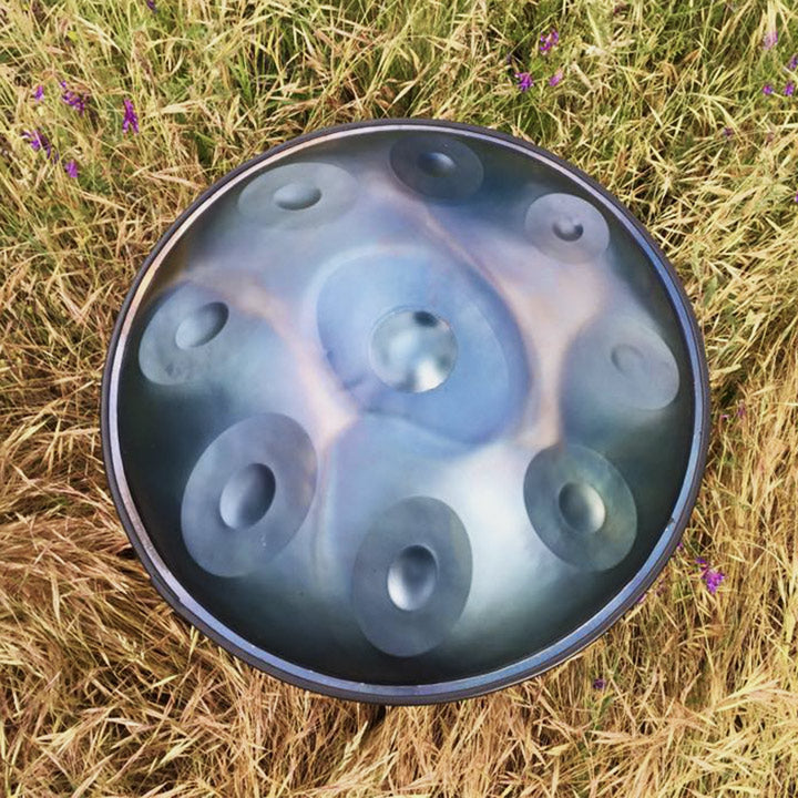 Nitrierte Mystical Handpan von oben mit einem silber-hellblauen Schimmer. Sie liegt auf einem gelben Feld mit lila Blumen.