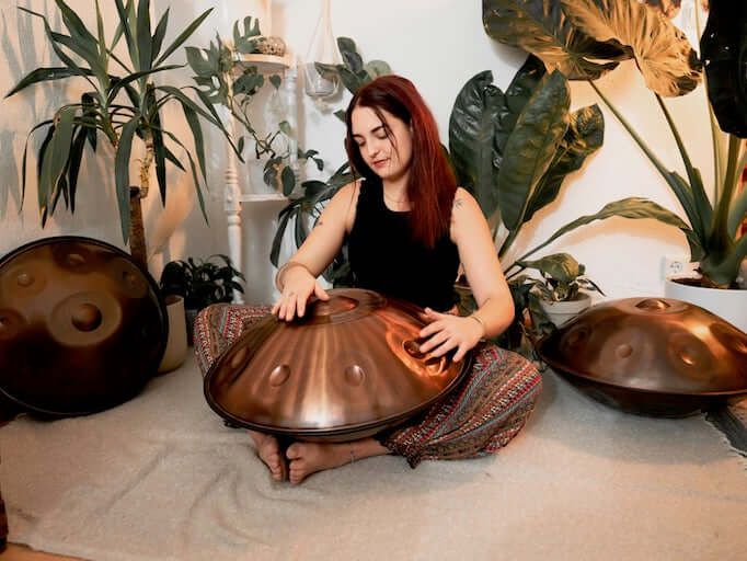 Handpanspielerin sitzt in einem schönen Raum mit Pflanzen und Handpans auf dem Boden und spielt eine AeloPan Handpan, die bronce glänzt