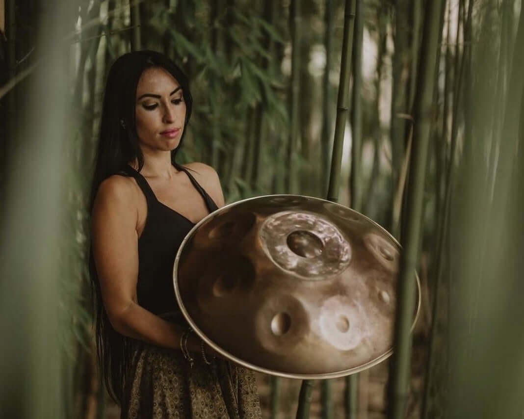 Eine Frau hält eine goldene Handpan zwischen mehreren Bambussträngen. Die Handpan spiegelt sich im Licht und schimmert dadurch silbern.