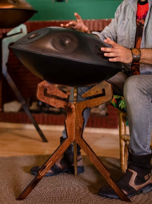 Schwarze, nitrierte Handpan steht auf einem hölzernen Katanui Sittingstand, welcher in Mitten eines gemütlichen, hellen Wohnzimmers steht, während ein Mann auf der Handpan spielt.