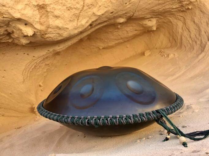 Nitrierte Handpan liegt auf einem sandigen Boden in einer Sandhöhle umrandet mit einem grün-grauen Schmuckband mit hölzernen Perlen am Ende.