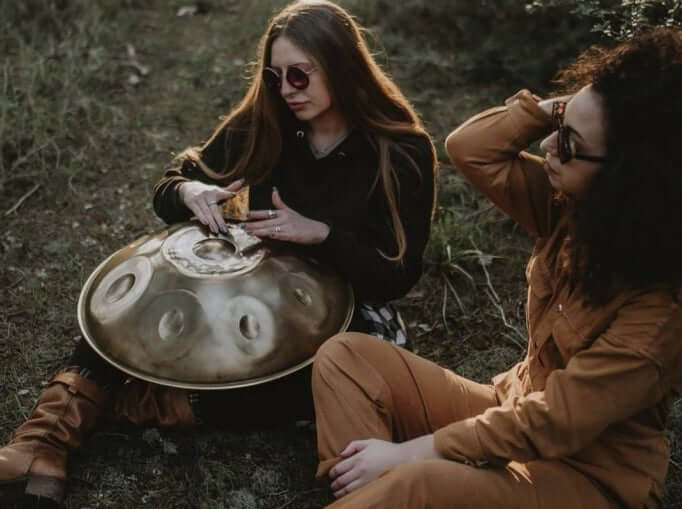 Zwei Frauen sitzen auf einer Wiese, während die eine auf einer goldenen, silberschimmernden Handpan spielt. Diese Handpan, sowie viele weitere kann man online im Handpan.World Store kaufen.